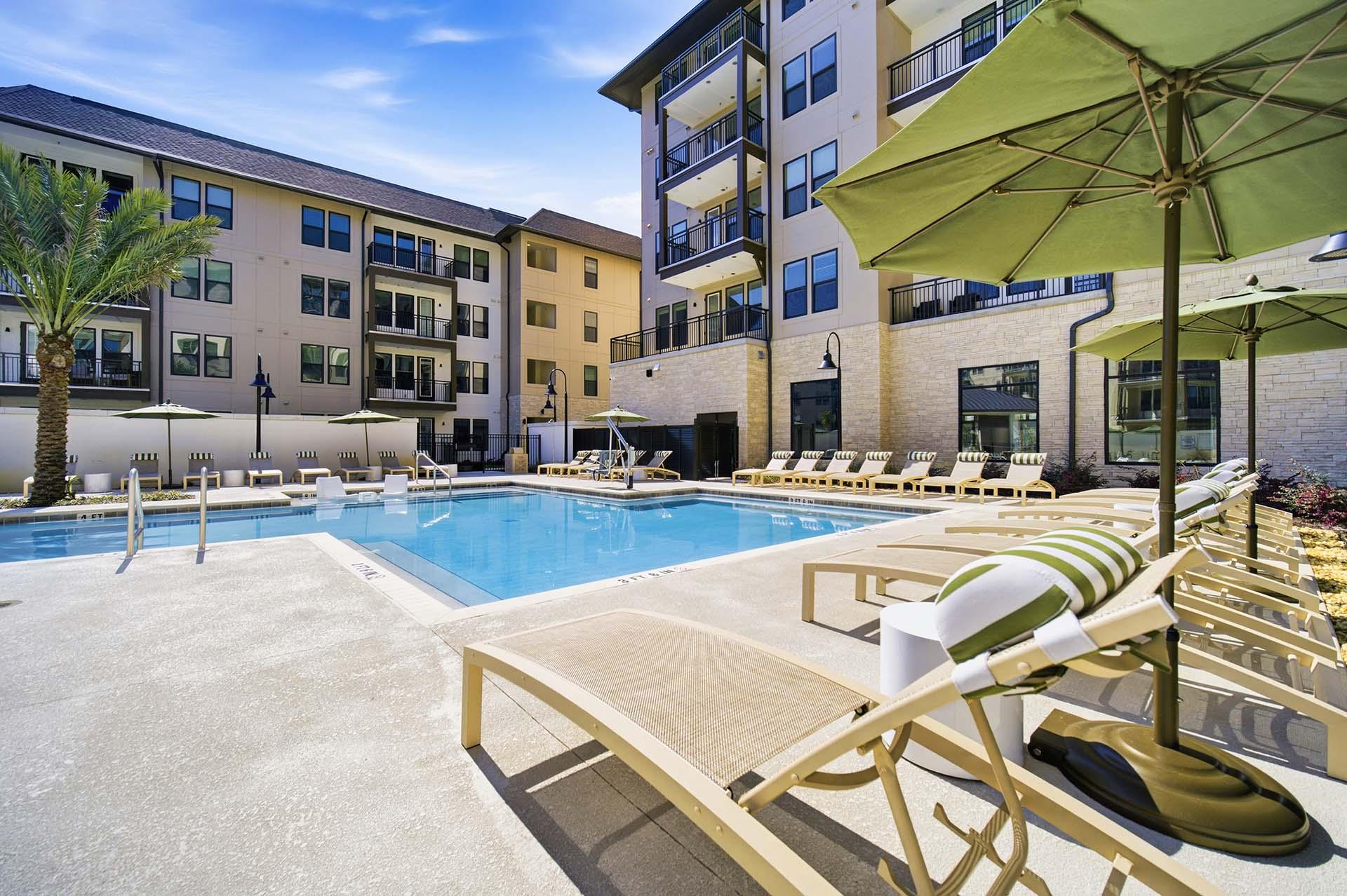 A pool area with sun loungers and umbrellas in front of apartment buildings.