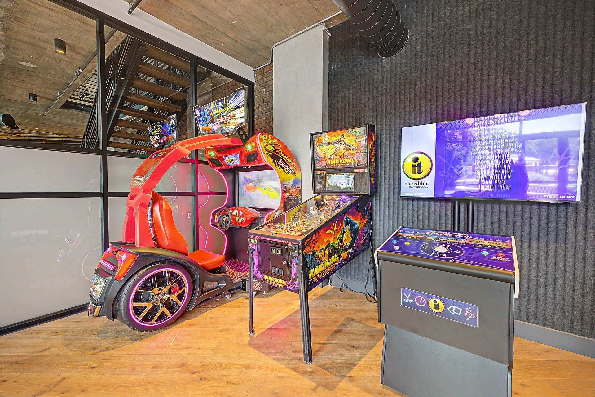A red and black car-themed arcade game machine sits in a room with a wooden floor and a grey trash bin.