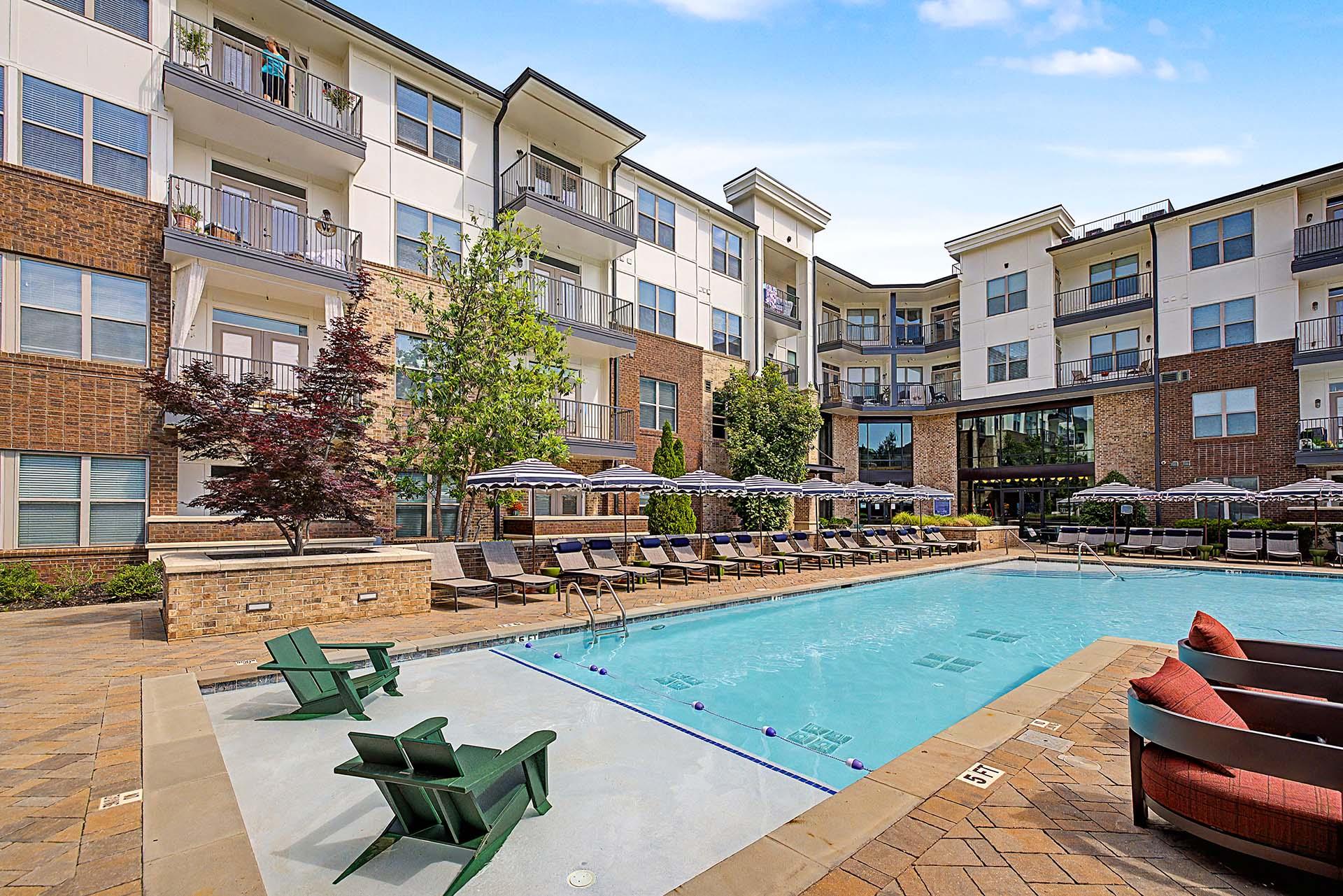 A large swimming pool surrounded by lounge chairs and umbrellas in front of apartment buildings.