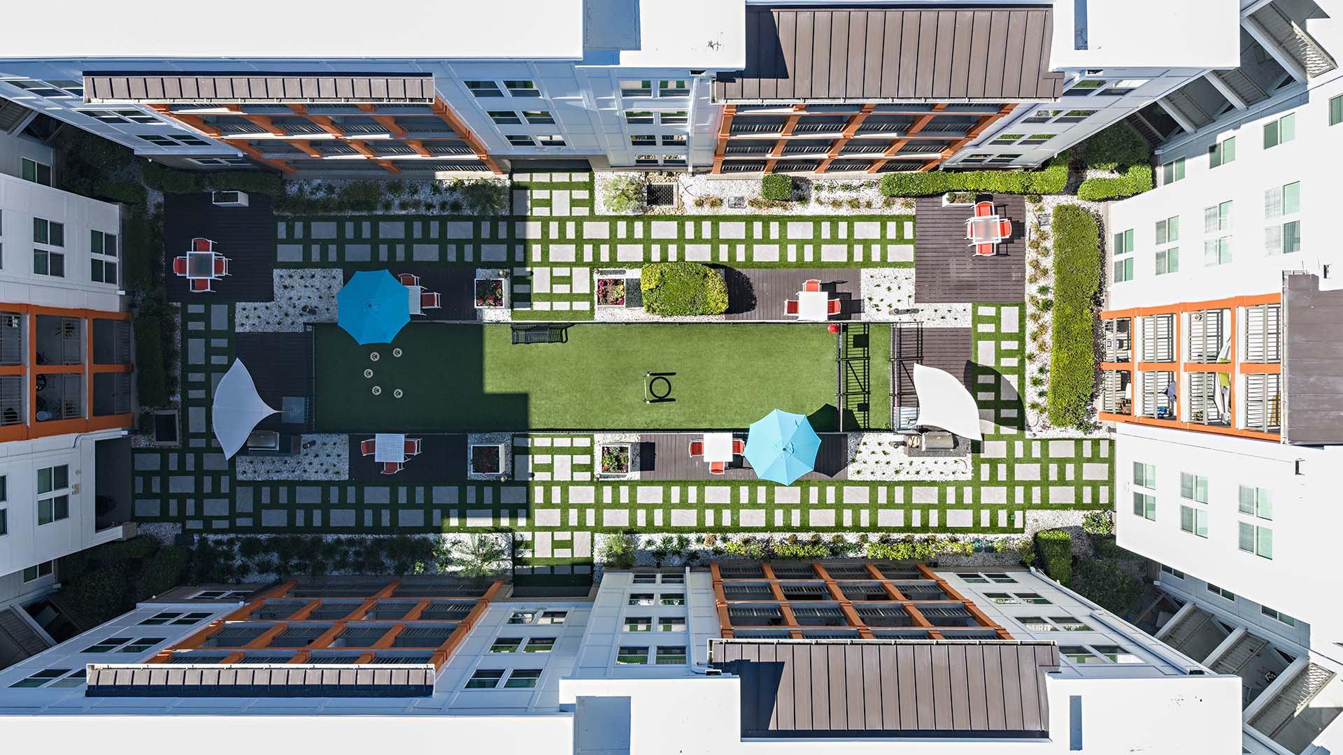 A top-down view of a courtyard surrounded by buildings with a green lawn and a blue umbrella.