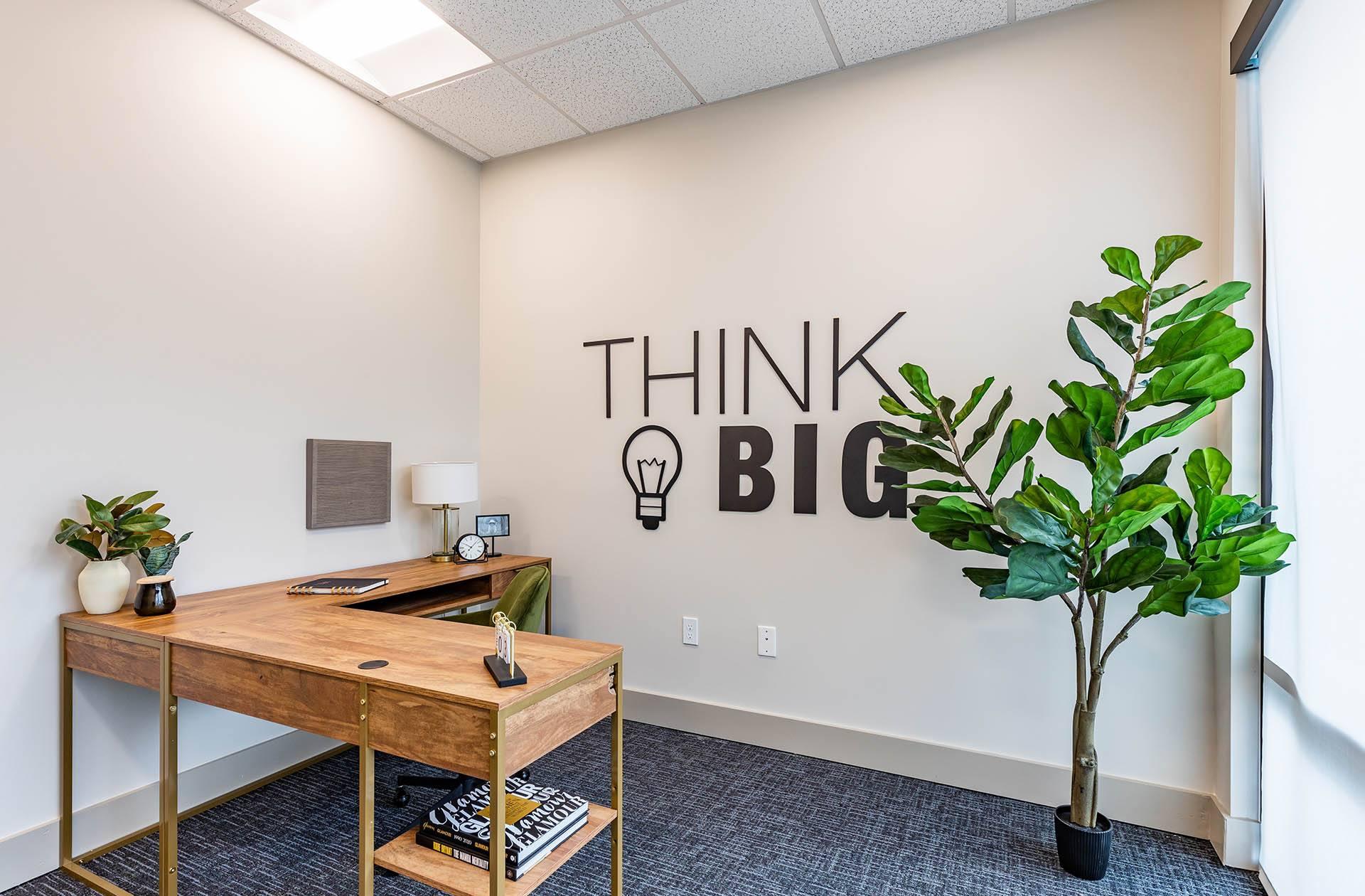 A room with a wooden desk and a plant with the words "Think Big" on the wall.
