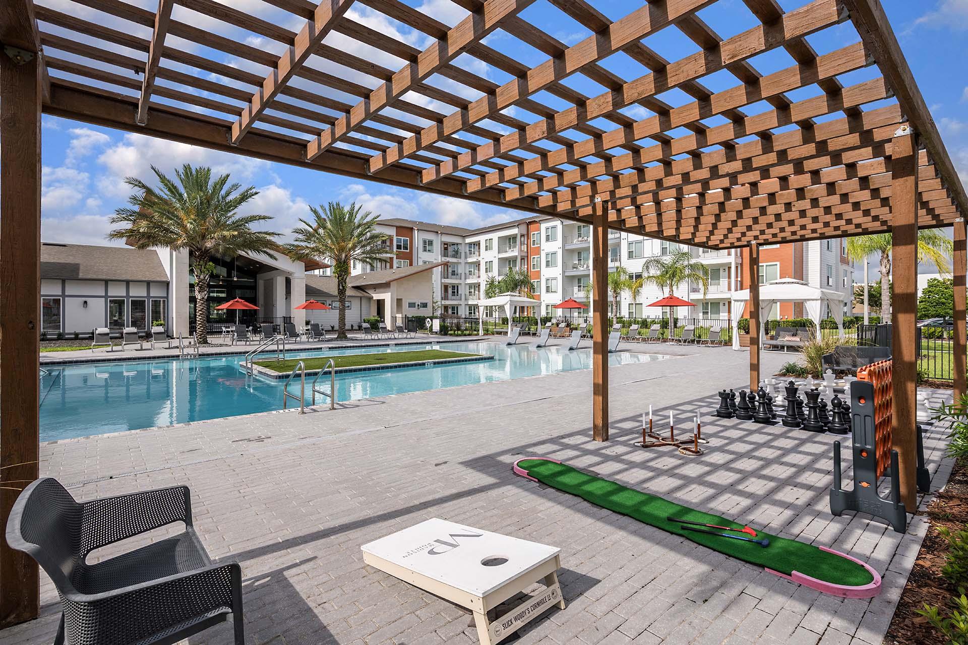 A wooden pergola over a poolside area with a lounge chair and a table.