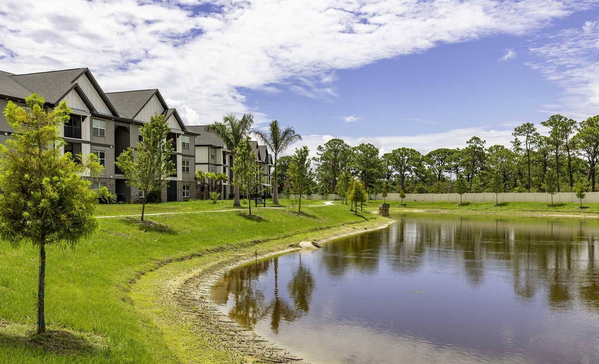 A serene landscape with a pond, a tree, and a row of houses.