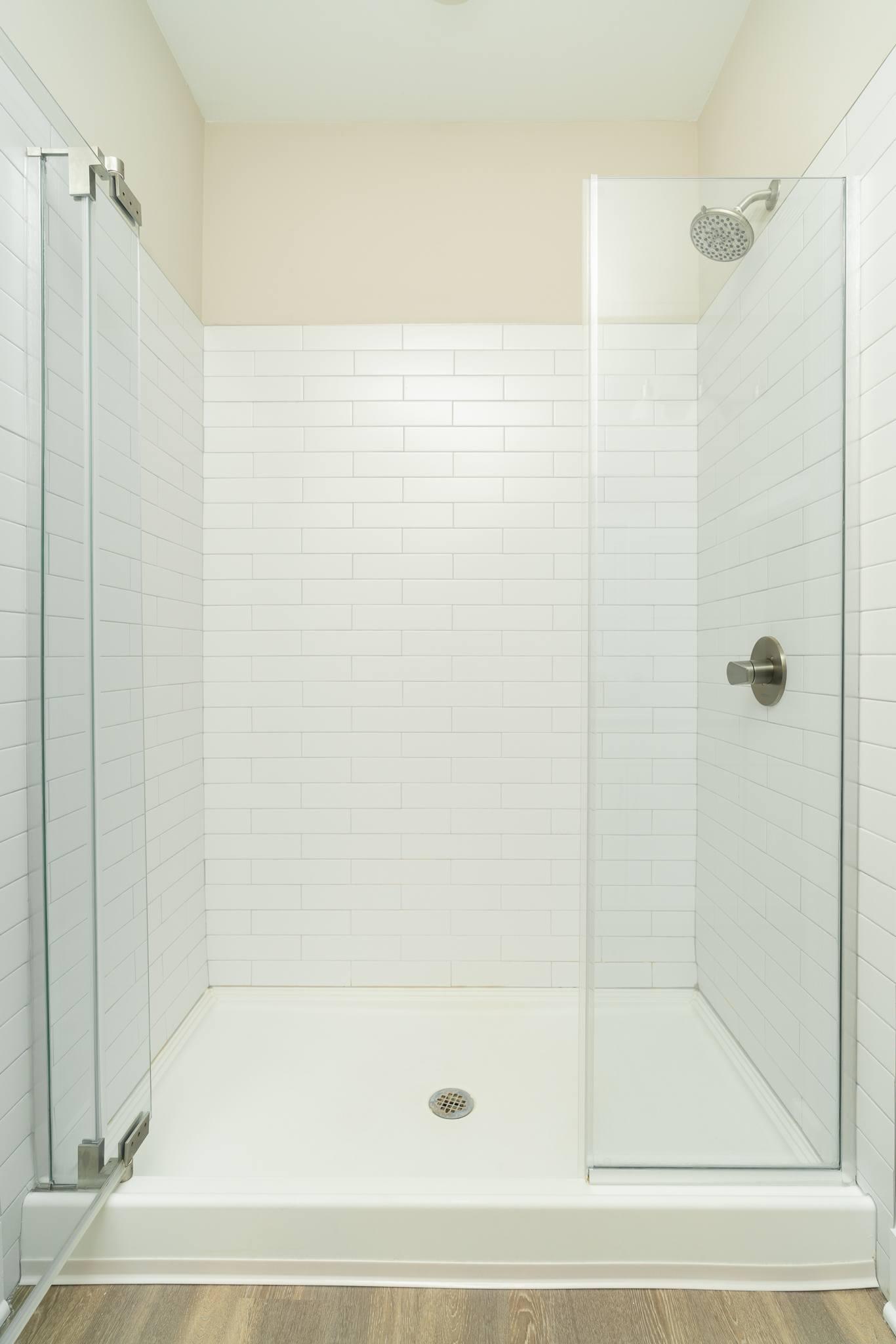 A white tiled shower with a glass door and a drain.
