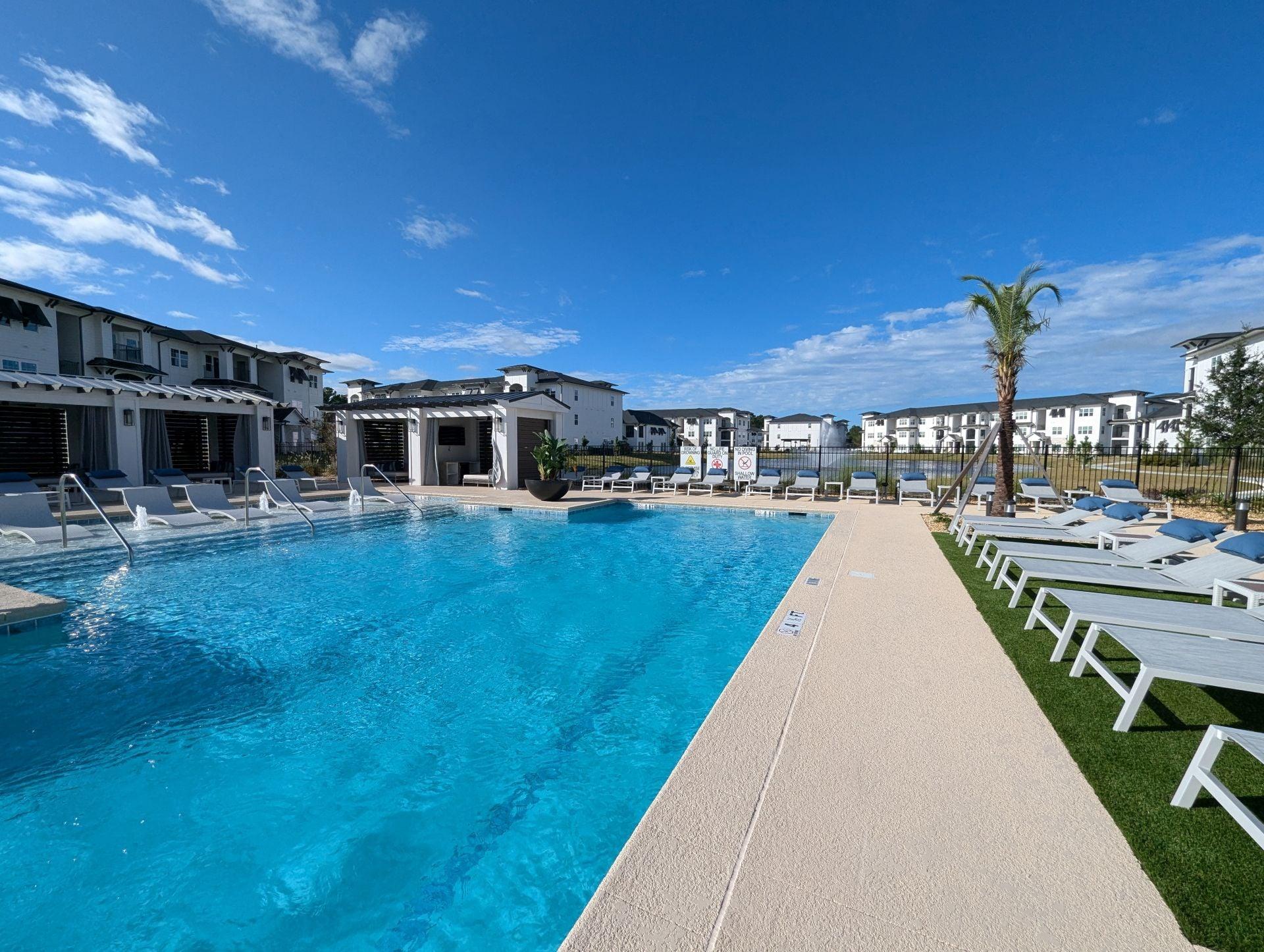 A large swimming pool with a clear blue water surrounded by white lounge chairs and buildings in the background.